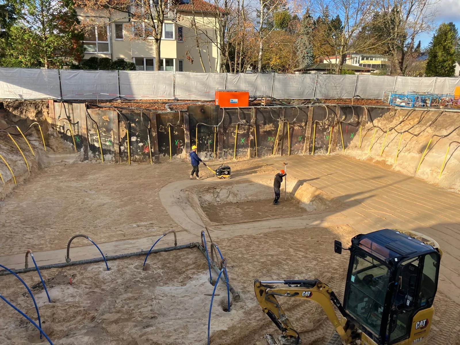 Fortgeschrittene Baugrube mit Trägerbohlwand, Vibrationsplatte und Mini-Bagger beim lagenweisen Aushub